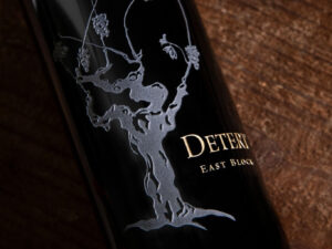 Detert East Block Cabernet Franc 2018 - OWC 3 Pack; Rectangular label featuring a dark bottle with a white grapevine and gold text, lying on a dark wood surface