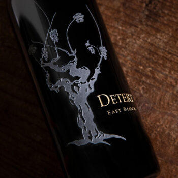 Detert East Block Cabernet Franc 2018 - OWC 3 Pack; Rectangular label featuring a dark bottle with a white grapevine and gold text, lying on a dark wood surface