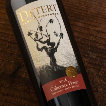 Detert Cabernet Franc Oakville 2018; Rectangular label showing a bottle of the wine lying on a dark wood surface. The bottle has a label that is beige featuring a dark brown grape vine. Text is gold, and gold on deep red.