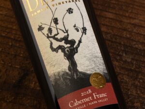 Detert Cabernet Franc Oakville 2018; Rectangular label showing a bottle of the wine lying on a dark wood surface. The bottle has a label that is beige featuring a dark brown grape vine. Text is gold, and gold on deep red.