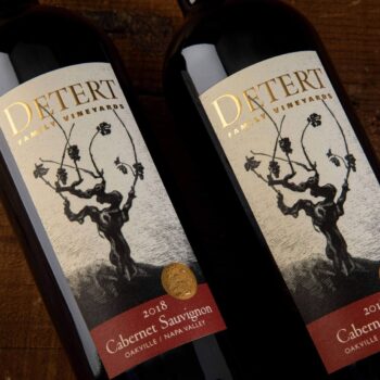 Detert Cabernet Sauvignon Oakville 2018; Two bottles of wine lying on a dark wood surface. The bottles have a label that is beige featuring a dark brown grape vine. Text is gold, and gold on deep red.