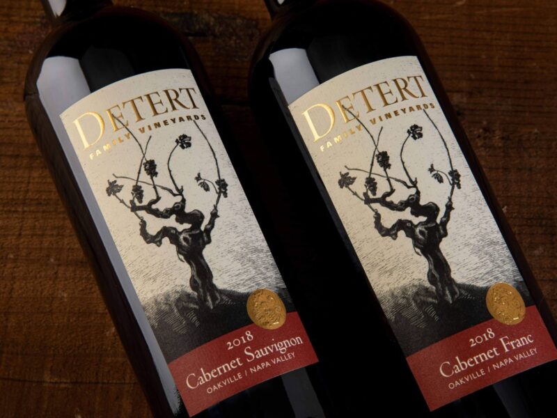Detert Cabernet Sauvignon Oakville 2018; Two bottles of wine lying on a dark wood surface. The bottles have a label that is beige featuring a dark brown grape vine. Text is gold, and gold on deep red.