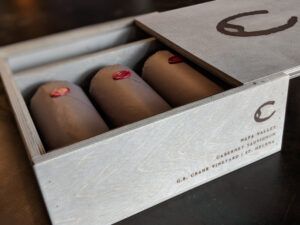 G.B. Crane Vineyard Cabernet Sauvignon 2017 OWC 3 Pack; Rectangular label , dark brown merging with black. Features a grey box, with the lid pulled back revealing three bottles wrapped in brown paper, with round, red wax seals