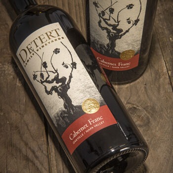 Detert Cabernet Franc 2014 3 Liter; A rectangular label showing two bottles of wine, one standing, one lying down, on a dark wood surface. The bottles have a label that is beige featuring a dark brown grape vine. Text is gold, and gold on deep red.