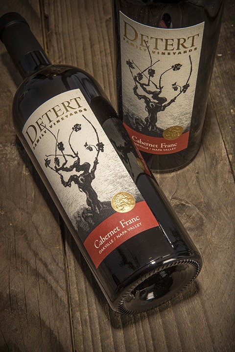 Detert Cabernet Franc 2014 3 Liter; A rectangular label showing two bottles of wine, one standing, one lying down, on a dark wood surface. The bottles have a label that is beige featuring a dark brown grape vine. Text is gold, and gold on deep red.