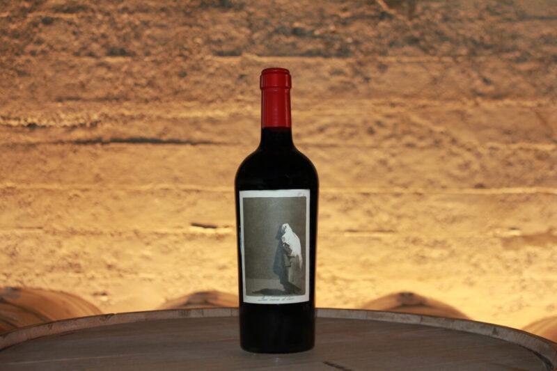 el Coco 2019; An oblong label showing a bottle of wine standing on a barrel top in the foreground, with more barrels beyond, against a sun kissed brick wall